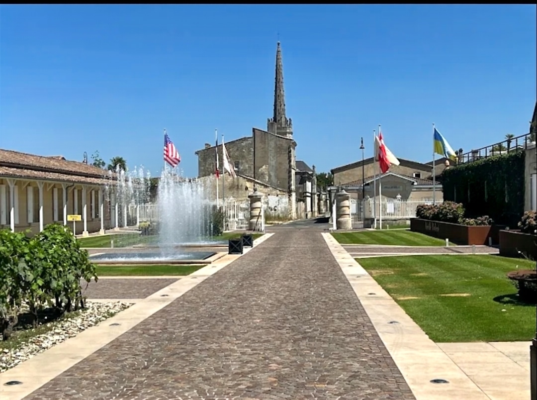 Allée pavée vers un clocher à Saint-Émilion, fontaines, drapeaux et vignes sous un ciel bleu.