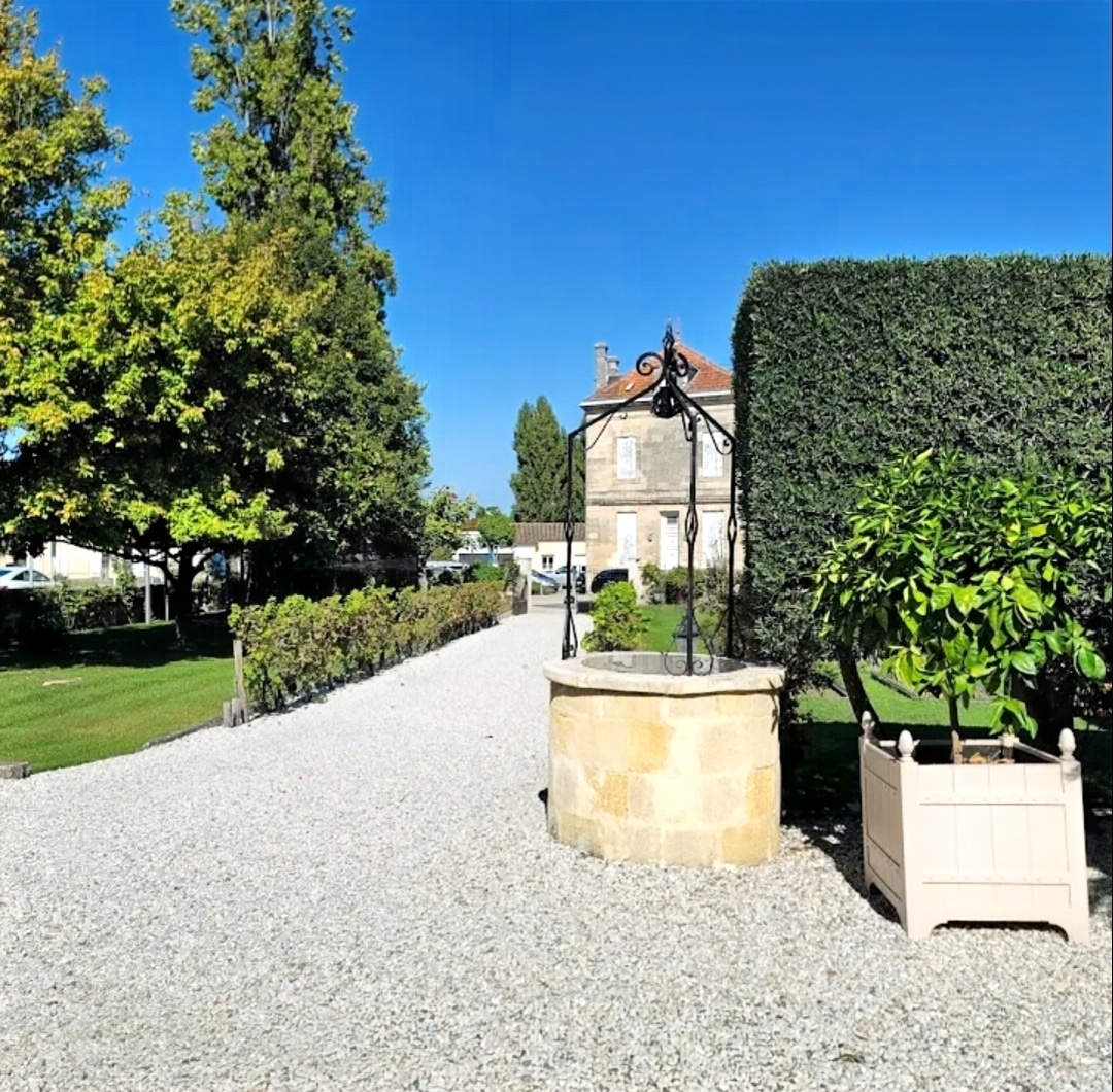 Puits traditionnel en pierre sur allée de gravier blanc devant une demeure ancienne sous un ciel bleu.